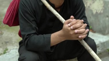 Elinde tahta bir sopayla yerde oturan siyah kıyafetli bir adam. Birey derin derin düşünür, elleri birbirine kenetlenir. Yakınlarda kırmızı bir çanta var..