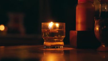 Cam tutacağında parlayan bir mum, sıcak ışık saçan ve karanlık bir yüzeye yansıyan. Arkaplanda bulanık ışıklar ve nesneler var, rahat bir atmosfer yaratıyorlar..