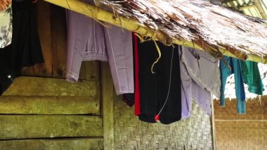 Sazdan çatıda asılı elbiseler, çeşitli renkler ve stiller sergileniyor. Arkaplanda ahşap bir duvar ve kırsal veya geleneksel yapılarda örülmüş bir doku yer alıyor..