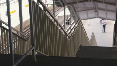 Beyaz korkuluklu bir merdiven manzarası, bir tren istasyonunda aşağı doğru iniyor. Arka plan platformda duran bulanık bir figürü gösteriyor, tren rayları görülebilir..