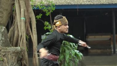 Dövüş sanatlarını doğal ortamında icra eden, geleneksel kıyafetler takan, saç bandı takan ve kuşak takan biri. Arkaplanda verimli yeşillik ve kırsal bir bina yer alıyor..