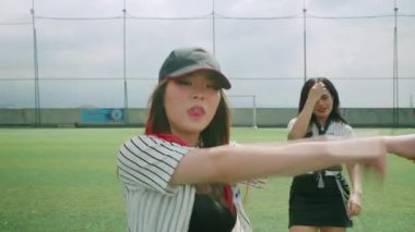 Spor sahasında iki genç bayanın dans ettiği canlı bir sahne. Beyzbol şapkalı ve çizgili gömlekli bir kadın enerjik bir şekilde hareket ederken, diğeri etek giyip arka planda dans ediyor. Ortalık aydınlık ve güneşli.