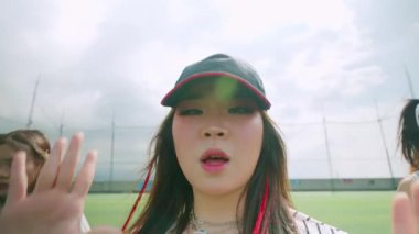 Spor sahasında enerjik dans eden genç kadınların olduğu dinamik bir sahne. Ön planda kep ve katmanlı mücevher takan bir kadın ve arka planda başkaları var. Gökyüzü parlak ve bulutlu, canlı atmosfere ekleniyor.