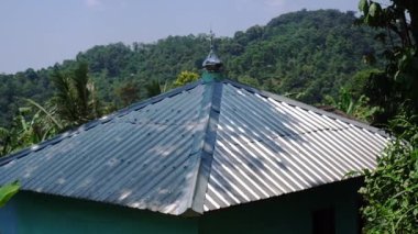 Arka planda yemyeşil ve tepelerle çevrili bir evin metal çatısının yakın görüntüsü. Çatıda güneş ışığını yansıtan dekoratif bir son var..