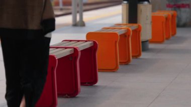 Tren istasyonunda kırmızı bir taburede oturan biri, arka planda turuncu tabureler sıralanmış. Şehir merkezi, oturma düzenine odaklı modern bir tasarıma sahip..