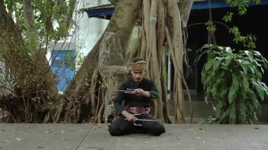 Geleneksel giyinmiş bir dövüş sanatçısı ağaçlar ve yeşilliklerle çevrili doğal bir ortamda silahlarla poz verir..