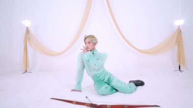 Zarif bir şahıs beyaz bir arkaplanda zarif bir şekilde poz verir. Nane yeşili bir elbise ve düz bir üst ve geniş bacaklı bir pantolon giyer. Sahne, arka planda kumaşla hafifçe aydınlatılıyor..