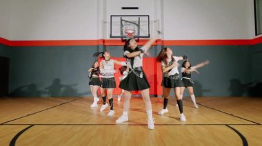 Bir grup dansçı bir basketbol sahasında, şık kıyafetlerle senkronize hareketler sergiliyorlar. Arka planda basketbol potası ve ahşap zemin var..