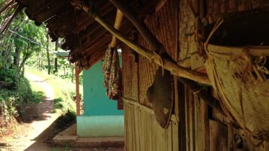 Yeşilliklerle çevrili sazdan çatılı ahşap bir evin yer aldığı köy manzarası. Geleneksel örülmüş eşyalar bambu direğine asılır ve toprak yol da mesafeye çıkar..