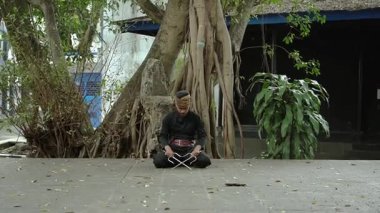 Geleneksel giyinmiş bir adam taş bir yüzeyde bağdaş kurmuş, elinde iki metal nesne tutuyor. Etrafı yemyeşil ve büyük bir ağaçla çevrili. Huzurlu bir atmosfer yaratıyor..