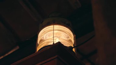 Kaliteli bir fener, ahşap bir kirişe asılı, sıcak bir parıltı saçıyor. Fenerin cam bir gövdesi ve metal bir çerçevesi var. Görünür örümcek ağları var. Bu da eski, kırsal bir ayarı gösteriyor..