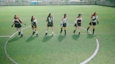 Futbol sahasında dans eden altı genç kadından oluşan bir grup. Sportif kıyafetler giyiyorlar, enerji ve coşku gösteriyorlar. Arka planda yeşil çim ve kale direkleri görünüyor..