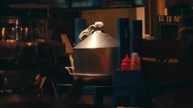 Loş bir restoranda mavi ahşap bir masada büyük metal bir tencere duruyor. Tencere kapakla kaplanmış ve üzerine kumaş örtülmüş. Yakınlarda ketçap ve hardal da dahil olmak üzere sos şişeleri var..