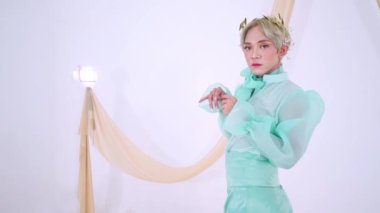 Şık bir nane yeşili elbise giyen, kabarık kollu, açık bir arkaplanda kumaşla zarif poz veren biri. Görünüşü, çiçekli bir başlıkla tamamlanmış..