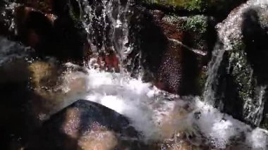 Kayaların üzerinde çağlayan suyun yakın çekim görüntüsü kabarcıklar ve sıçramalar yaratıyor. Güneş ışığı sudan yansıyarak taşların dokusunu ve suyun hareketini vurgular..