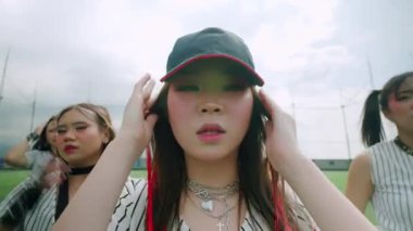 Bir grup genç bayan dışarıda dans gösterisi yapıyor, beyzbol temalı spor kıyafetleri giyiyorlar. Ön plandaki bir kadının saçlarında kırmızı röfleler var ve enerjik bir ifade sergiliyor..