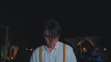 Koyu renk saçlı ve ciddi bir yüzü olan beyaz pantolon askısı takmış genç bir adam, loş bir açık hava ortamında duruyor. Arka planda yumuşak ışıklandırma ve dekoratif elementler var. Karamsar bir atmosfer yaratıyorlar..