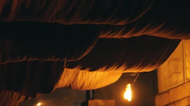 Perdeli kumaş tavanın altında asılı bir ampulün olduğu loş bir sahne. Ampulün sıcak parıltısı, koyu renkli kumaşla çelişiyor ve rahat bir atmosfer yaratıyor..