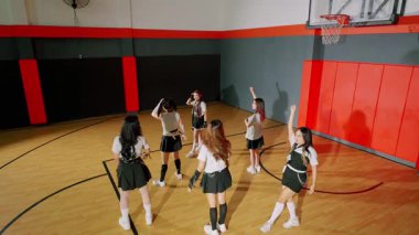 Bir basketbol salonunda dans rutini uygulayan bir grup uyumlu kıyafetli kız. Sahne, hareketlerini arkadan yakalıyor. Salonun ahşap zeminini ve basketbol potasını gösteriyor..