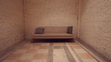 İki gri yastıklı bej bir kanepenin dokulu beyaz tuğla bir duvara dayandığı minimalist bir iç mekan. Zemin, aydınlık ve koyu fayanslı desenli bir tasarıma sahip, rahat bir atmosfer yaratıyor..