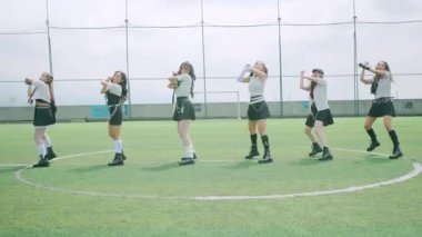 Yeşil bir futbol sahasında, şık kıyafetler giymiş altı dansçıdan oluşan bir grup. Harekete geçtiler, enerjik ifadelerle koreografi sergiliyorlar. Arka planda açık bir gökyüzü ve spor alanı unsurları yer alıyor.