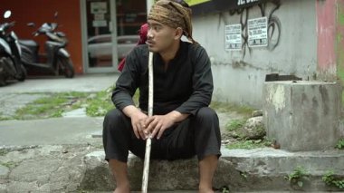 Bir taş basamakta oturan bir adam, geleneksel siyah giysiler ve türban takmış, elinde tahta bir sopa tutuyor. Şehir unsurları ve park etmiş motosikletlerle derin derin düşünüyor gibi görünüyor..