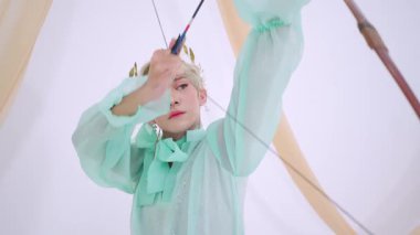 Nane yeşili kıyafetli biri ok ve yayla nişan alır. Odaklanma ve kararlılık gösterir. Arka planda yumuşak perdeler var, sahneye tuhaf bir dokunuş ekliyor..