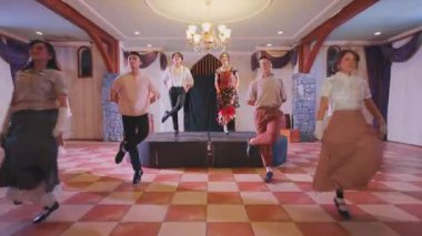 Güzel dekore edilmiş bir salonda bir grup dansçı sahnede gösteri yapıyor. Dansçılar klasik kıyafetler giyiyorlar, canlı bir performans sergiliyorlar. Ayarda zarif bir ışıklandırma ve bir avize var..