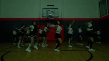 Bir grup dansçı basketbol potasıyla bir spor salonunda gösteri yapıyor. Aynı kıyafetleri giyiyorlar, senkronize hareketleri ve enerjik ifadeleri sergiliyorlar. Işık çarpıcı bir atmosfer yaratıyor..
