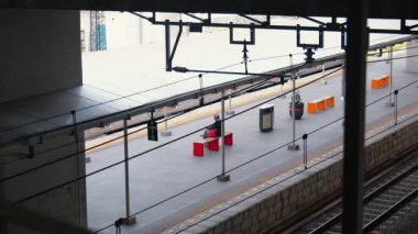 Kırmızı ve turuncu renkli oturma düzenlerinin yer aldığı bir tren istasyonu platformu manzarası. Platform boş, birkaç genel bina ve raylar görülebilir..