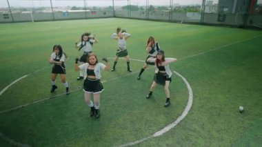 Futbol sahasında dans eden altı genç kadından oluşan bir grup. Etek ve botlarla uyumlu kıyafetler giyiyorlar, enerjik hareketler sergiliyorlar. Arkaplanda yeşil bir çim sahası ve şehrin ufuk çizgisi yer alıyor..