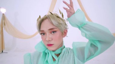 Kısa, açık renkli saçları olan, zarif, nane yeşili bir bluz giyen biri. Hassas bir başlıkları var ve arka planda kumaşla örtülü yumuşak, sanatsal bir ortamda poz veriyorlar..