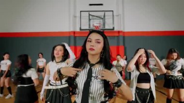 Bir spor salonundaki dinamik dans gösterisinde şık kıyafetler giymiş bir grup genç kadın olacak. Merkezi dansçı, elleri başının üstünde, koreografi eşgüdümlü bir şekilde etrafını sarmış duruyor..