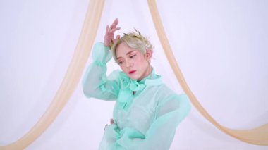 Açık yeşil giysili, kabarık kollu bir insan yumuşak, pastel bir arka plana karşı zarifçe poz verir. Zarif bir başlıkları ve sakin bir ifadeleri var..