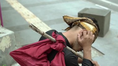 Başörtüsü takan ve omzunun üzerine kırmızı bir bez torba asılmış, kafası aşağıda oturan, dalgın ya da sıkıntılı görünen bir kişi..