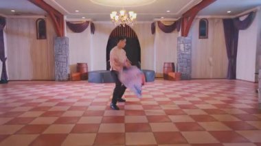 Geniş bir salonda, ahşap kirişler ve avizelerle canlı bir dans gösterisi. Bir dansçı renkli bir bayrak tutarken, diğerleri geleneksel kıyafetlerini giyerek canlı bir atmosfer yaratıyor..