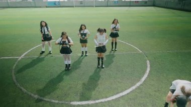 Şık giyinmiş altı genç kadından oluşan bir grup yeşil bir spor sahasında daireler çiziyor. Siyah beyaz giyinmişler, etek ve şort karışımı, modaya uygun, sportif bir görünüm sergiliyorlar. Güneş ışığı çimenlerin üzerinde uzun gölgeler bırakır.