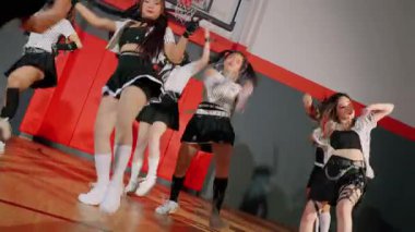 Bir grup dansçı bir spor salonunda koreografi gösterisi yapıyor. Siyah ve beyaz karışımı şık kıyafetler giyiyorlar, dinamik hareketler ve ifadeler sergiliyorlar. Kurulumda ahşap zemin ve renkli duvarlar yer alıyor..