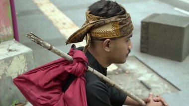 Geleneksel bir saç bandı ve siyah gömlek takan genç bir adam omzunda bir sopayla oturuyor, kırmızı bir bezle. Arka plan kaldırımlı bir şehir caddesini gösteriyor..