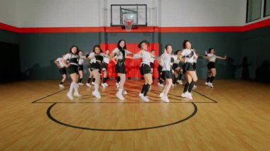 Bir grup dansçı bir basketbol salonunda koreografi gösterisi yapıyor. Aynı kıyafetleri giyiyorlar ve enerjik hareketler sergiliyorlar. Spor salonunda basketbol potası ve ahşap zemin var..
