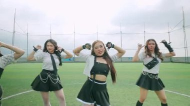 Çimenli bir alanda dans eden, çizgili ve aksesuarlı şık kıyafetler giyen bir grup genç kadın. Atmosfer canlı ve enerjiktir. Arka planda bulutlu bir gökyüzü vardır..