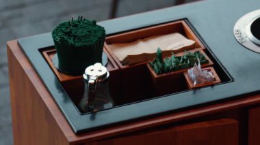 İçinde çeşitli çeşniler ve çatal bıçaklar bulunan siyah yüzeyi olan ahşap bir masa. Yeşil yemek çubukları, peçeteler ve içinde bitkiler ve şeker olan küçük kaplar var. Cam bir kavanoz da var..