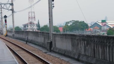 Arka planda beton bir duvar ve yeşillik ile çevrili raylı bir demiryolu platformu manzarası. Olay yeri biraz puslu, nemli bir atmosferi işaret ediyor..