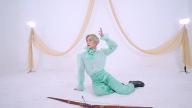 Zarif bir şahıs beyaz bir arka planda zarif bir şekilde poz verir, büyük bir papyon ve nane yeşili bir elbise giyer. Sahne, arka planda kumaşla hafifçe aydınlatılıyor ve zarif bir atmosfer yaratıyor..