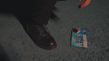 Mavi desenli bir yüzey üzerinde renkli bir biletin yanında duran siyah bir ayakkabının yakın plan çekimi. Bilette 'TICKET' kelimesi belirgin bir şekilde gösterilmiş, bir olay veya çekim için olduğu öne sürülüyor.