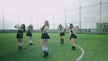 Bir futbol sahasında altı dansçıdan oluşan bir grup, şık siyah beyaz kıyafetler giymişler. Canlı bir atmosferle senkronize hareketleri sergiliyorlar..