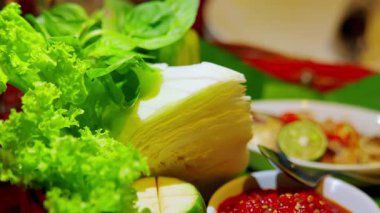 Marul, lahana ve kireç de dahil olmak üzere canlı bir sebze gösterisi, yanında bir kase baharatlı sos ve bir tabak erişte, yeşil yapraklı arka planda..