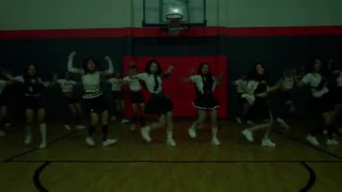 Bir grup ponpon kız spor salonunda senkronize dans gösterisi yapıyor. Aynı üniformayı giyiyorlar ve enerjik, gösterişli takım çalışması ve coşkuları var..