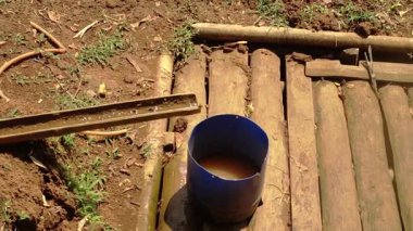 Kahverengi suyla dolu mavi bir kova toprak ve yeşil çimlerle çevrili ahşap bir yüzeyde oturur. Yakınlarda su için küçük bir kanalı olan bambu benzeri bir yapı görülebilir..