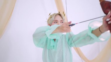 Açık yeşil kıyafetli, ok ve yayla dikkatlice nişan alan biri. Arka planda yumuşak perdeler var, dingin bir atmosfer yaratıyor. Bireyin çiçekli bir tacı ve odaklanmış bir ifadesi var..